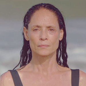 A woman with wet, dark hair stands in front of a blurry beach background, looking directly at the camera with a calm, neutral expression. She is wearing a black swimsuit.