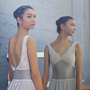 A ballerina in a light gray leotard and bun hairstyle stands in front of a mirror, looking over her shoulder at her reflection in a softly lit studio.