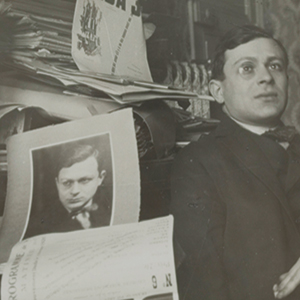 A man in a suit sits beside a cluttered desk stacked with papers and a large photograph of himself. The background is filled with more papers and printed documents.