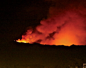 A large wildfire burns brightly on a hillside at night, with thick smoke rising into the sky. Below, city lights and illuminated buildings are visible in the foreground.