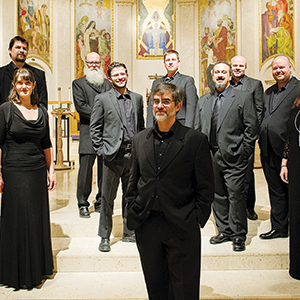 A group of ten people dressed in formal black attire stands in a church with ornate paintings and arches in the background, posing together and smiling for the photo.