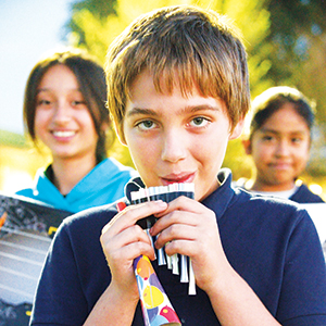 A boy in a navy blue shirt plays a pan flute made from straws, while two girls smile in the background outdoors on a sunny day.