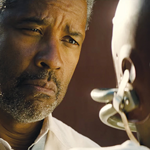 A man with a serious expression looks intently at someone wearing a metal restraint around their mouth and neck, set against a blurred, reddish-brown background.