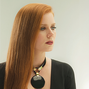 A woman with straight red hair, wearing a black top and a chunky black and gold necklace, looks to the side against a light, plain background.