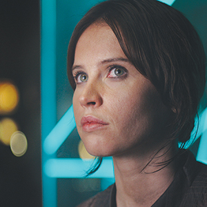 A woman with short brown hair and light eyes looks up thoughtfully. She is illuminated by soft, blue neon lights in the background, giving the scene a futuristic feel.