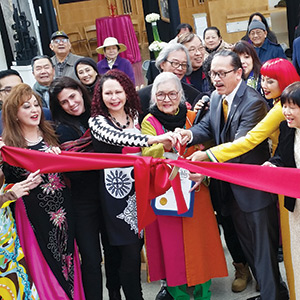 A diverse group of people, smiling and gathered closely, cut a large red ribbon together at an indoor event, celebrating an opening or special occasion. Some wear colorful, festive clothing.
