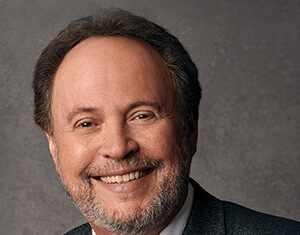 A middle-aged man with short brown hair and a beard is smiling. He is wearing a gray suit jacket, white shirt, and a black tie, with a neutral gray background behind him.