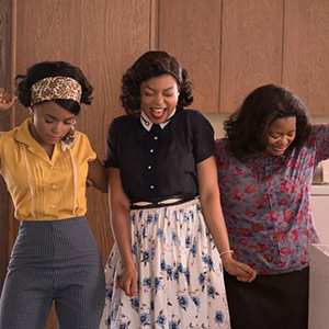 Three women stand close together indoors, smiling and holding hands. One wears a yellow blouse and headband, the second wears a dark top and floral skirt, and the third wears a floral shirt. They appear joyful and celebratory.