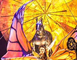 A performer in a butterfly costume kneels on stage, with large orange wings spread open and a large animal statue with a glowing yellow background behind her.
