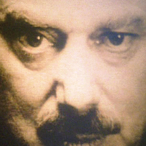 A close-up view of a mans intense face, focusing on his serious eyes and mustache, with a grainy, sepia-toned effect.