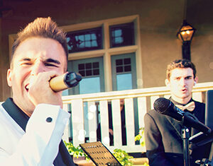 A man in a white suit sings passionately into a microphone, while another man in a black suit plays a keyboard outdoors in front of a building with windows and a railing.