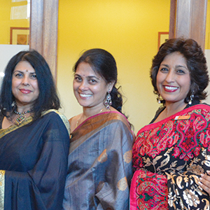 Three women in colorful sarees stand together, smiling for the camera in an indoor setting with a yellow background.