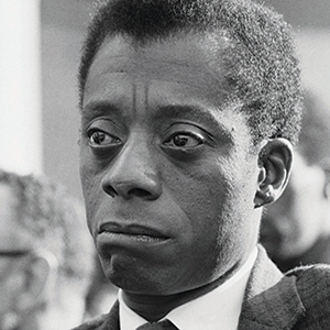Black-and-white close-up of a man with short hair and a serious expression, wearing a suit and tie. He looks slightly to the side, and people are blurred in the background.