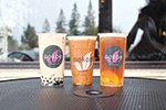Three bubble tea drinks in plastic cups with colorful logos are placed on an outdoor table. The drinks vary in color and ingredients, and blurred trees and a person are visible in the background.