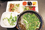 A bowl of clear soup with green onions, a plate of sliced meat with rice noodles, and a trio of dipping sauces arranged neatly on a table.