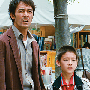 A man wearing a brown jacket stands next to a boy in a white jacket. They are outdoors near a tree and market stalls with books in the background, both looking intently at something off-camera.