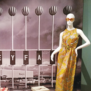 A mannequin in a colorful, patterned dress and sunglasses stands in front of a black-and-white vintage photo backdrop showing a Macy’s store and parked cars.
