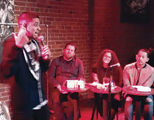 A man stands with a microphone performing on stage in front of a brick wall, while three people sit at a table listening and watching him. The setting appears to be a comedy club or small venue.