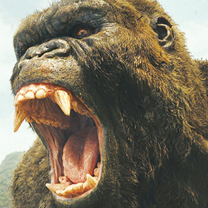 A close-up of a giant gorilla roaring with its mouth wide open, showing sharp teeth and fangs. The creature has brown fur and a fierce expression.