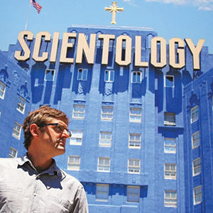 A man in glasses stands in front of a large blue building with a sign reading SCIENTOLOGY in bold letters and a gold cross on top. An American flag is visible against a blue sky in the background.