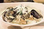 A bowl of creamy pasta topped with mussels, grated cheese, and fresh herbs, served in a white dish with a spoon on the side.