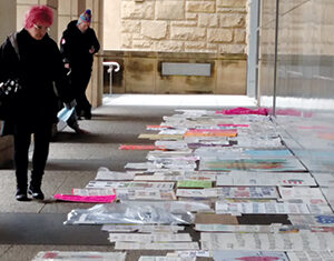 A person with pink hair walks past protest signs and posters laid out on the ground along a covered walkway beside a stone building. Another person stands further away, also looking at the signs.