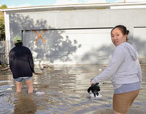 Two people wade through knee-deep floodwater in front of a garage with an orange X marked on the door. One person faces the camera while holding shoes; the other walks away. Debris floats in the water.