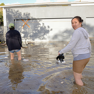 Two people wade through knee-deep floodwater in front of a garage with an orange X marked on the door. One person faces the camera while holding shoes; the other walks away. Debris floats in the water.