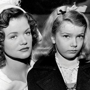 Black-and-white photo of a woman with wavy hair and a young girl with long hair and a bow. Both appear serious, looking slightly past the camera, dressed in vintage clothing.