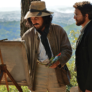 Two men stand outdoors; one, wearing a hat and casual clothing, paints on an easel while holding a palette. The other, dressed more formally, watches him. Trees and a blurred landscape are visible in the background.