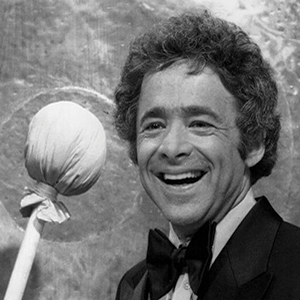 A smiling man in a tuxedo stands next to a large mallet and a gong in the background. The image is in black and white.