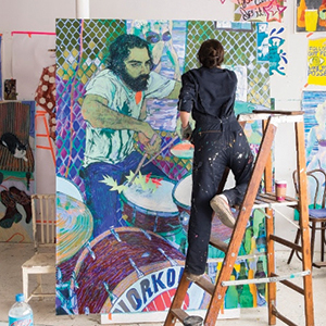 An artist in dark overalls stands on a ladder, painting a colorful portrait of a drummer on a large canvas in a brightly lit art studio filled with other artworks and supplies.