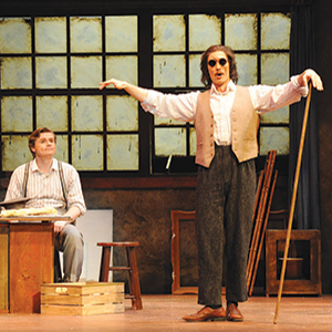 Two men perform on stage; one sits at a desk with papers, while the other stands wearing dark glasses and a vest, holding a cane and gesturing with his arms outstretched. A large window is behind them.