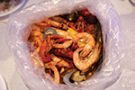 A clear plastic bag filled with cooked shrimp, mussels, corn, and sausage, seasoned and ready to eat, placed on a white surface.