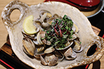 A ceramic dish filled with cooked clams, garnished with microgreens and a lemon wedge, served on a wooden board.