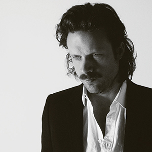 Black-and-white photo of a man with medium-length hair and a mustache, wearing a dark blazer over an unbuttoned white shirt, looking downward with a serious expression against a plain background.