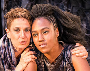 Two people sit close together, one with short hair and the other with long braided hair. The person with short hair has their arm around the other, both wearing textured, gray-toned clothing and looking ahead with serious expressions.