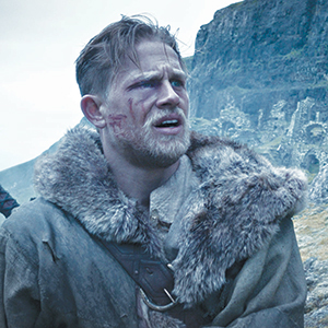 A rugged man with short blond hair and a beard, wearing a fur-lined cloak, stands outdoors with a rocky cliff in the background. His face is dirty and scratched, and he looks ahead with a concerned expression.