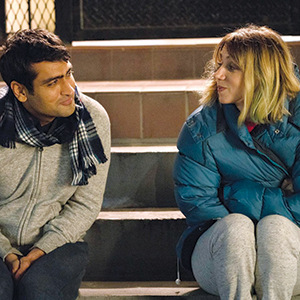 A man and a woman sit on outdoor steps at night, facing each other and smiling warmly. The man wears a scarf and light jacket, while the woman wears a blue puffy coat. They appear to be having a friendly conversation.