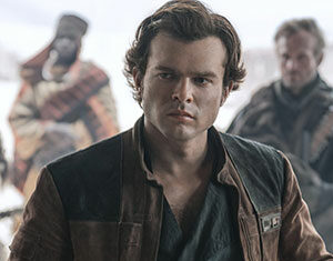 A young man with wavy brown hair wearing a brown and black jacket stands confidently, looking serious. Blurry figures in rugged clothing are visible in the background.