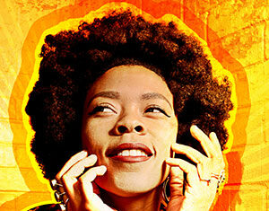 A woman with an afro hairstyle smiles and looks upward, her hands touching her face. She wears patterned clothing and rings, with a vibrant orange and yellow background behind her.