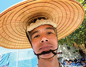 A man wearing a traditional straw hat and patterned shirt stands outdoors, looking at the camera. There are people, trees, and blue tents or structures in the background, suggesting a festival or outdoor event.