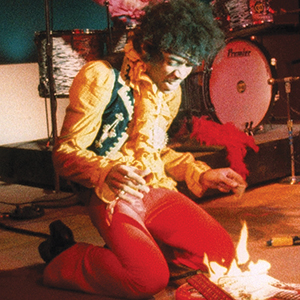 A musician in a yellow shirt and red pants kneels on stage, setting an electric guitar on fire, with drums visible in the background.