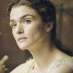 A woman with wavy brown hair pulled back looks slightly to the side with a calm, thoughtful expression. She wears a light-colored garment and is in a softly lit indoor setting.