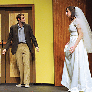 A man in a brown blazer and khaki pants stands facing a woman in a white wedding dress and veil on stage, both appearing to be in the middle of a conversation or dramatic moment.