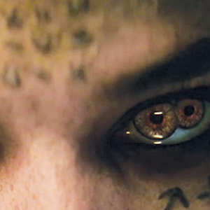 A close-up of a persons face showing intense, reddish-brown eyes with an unusual, double-pupil effect. The skin around the eyes has dark markings and patterns. The image has a mysterious and eerie atmosphere.