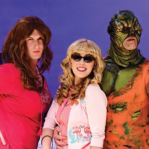 Three people posing together: one in a red dress and long wig, one in sunglasses with curly blonde hair and a pink outfit, and one in a green and orange monster costume with a distorted face.