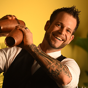 A smiling man with a beard, tattooed arm, and spiked hair shakes a cocktail shaker. He wears a white shirt and black vest against a yellow background with green plants.