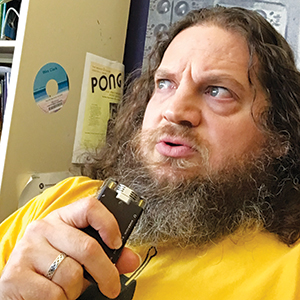 A bearded person with long hair, wearing a yellow shirt, holds a recording device and looks to the side. Behind them are a wall with papers, including one labeled PONG, and a blue label that resembles a CD.