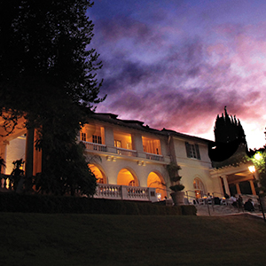 A large, elegant building with arched windows and balconies is warmly lit at dusk, set against a dramatic, colorful sky with trees framing the scene.